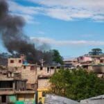 Imagen que muestra la devastación causada por pandillas en Haití, con viviendas incendiadas y humo en el barrio de Vivy Mitchel. En la imagen se observan residentes evacuando la zona en medio de la tensión y el caos, con personas llevando sus pertenencias y buscando refugio ante la violencia.