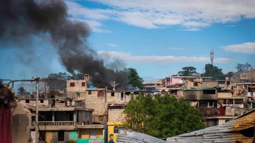 Imagen que muestra la devastación causada por pandillas en Haití, con viviendas incendiadas y humo en el barrio de Vivy Mitchel. En la imagen se observan residentes evacuando la zona en medio de la tensión y el caos, con personas llevando sus pertenencias y buscando refugio ante la violencia.