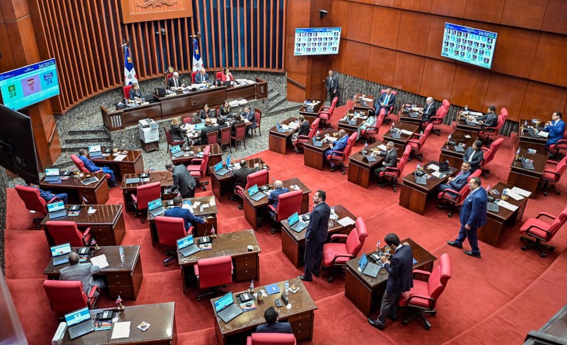 Reunión del Senado de la República Dominicana donde se discute y aprueba la modificación de la Ley del Consejo Nacional de la Magistratura, con enfoque en la adecuación de los artículos que regulan la selección del Procurador General y los jueces de altos tribunales. La imagen muestra a varios senadores en el salón de sesiones, destacando el ambiente de debate legislativo.